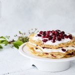 Grote ronde millefeuille met kersen en verse roomkaas. Deze staat op een witte taartschaal op een bleke houten tafel. Op tafel ligt ook een takje groen voor extra sfeer. 