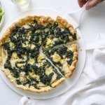 Op een taartschaal ligt de aangesneden quiche. Er is een witte taartschep onder één stukje quiche om naar een bordje te gaan. Langs de quiche staat ook nog een portie gemengde sla, voor nog een extra portie groenten. 