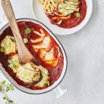 Pakjes van courgette liggen in witte ovenschaal met blauw randje. De tomatensaus ligt onder de ravioli van courgette, waardoor deze goed herkenbaar zijn. Er werd net een portie uitgeschept om meteen te eten. 