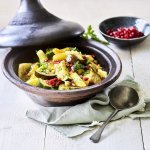 Er staat een tajine op tafel, de tajine heeft een zwarte kleur en is gevuld met stukjes aardappel, courgette, venkel, abrikozen, kikkererwten en granaatappelpitten. 