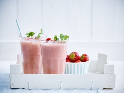 2 smoothies van aardbeien en melk. Ze staan in een houten bakje met een portie verse aardbeien er naast. De smoothies zijn afgewerkt met een takje koriander en kartonnen licht blauw rietje