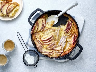 Een clafoutis van appelstukkjes staat op het aanrecht in een zwarte fonte schaal. Er is al een portie uitgeschept en staat op een bordje naar de pot. Er is ook bloemsuiker over het dessert gestrooid en de koffie is al uitgeschonken. 