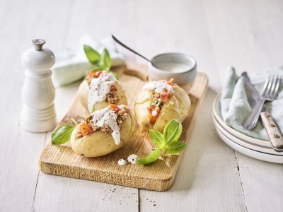 Enkele klaargemaakte en afgewerkte gepofte aardappelen liggen op een houten snijplank. Ze zijn gevuld met gehakt, stukjes tomaat en afgewerkt met een geitenkaasdip. Naast de aardappelen staan enkele bordjes, bestek en servetten om meteen te beginnen. 