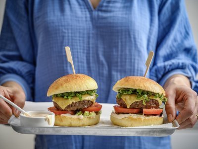 Een vrouw staat in beeld met haar romp draagt een plateau met hierop twee hamburgers. De hamburgerbroodjes hebben een mooi goudgele kleur, ze werden gemaakt met aardappelpuree in het deeg. Verder zijn de burger belgd met een schijfje vlees, kaas, tomaat, sla en saus natuurlijk. 