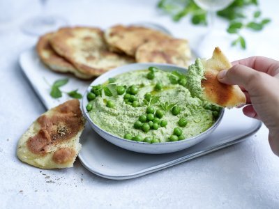 Krokant gebakken platbrood ligt op een serveerschaaltje waarbij een dip van erwten en roomkaas in geserveerd. De dip is gemixt en heeft een mooie groene kleur, om af te werken zijn er nog enkele verse erwtjes bovenop gestrooid. Een hand neem alvast een stukje platbrood om te dippen in gezonde groentedip. 