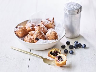 In een diep wit bord ligt een wit boterpapiertje met hierin enkele oliebollen. Er is eentje uitgeprikt en ligt op tafel doormidden gesneden, hier zie je duidelijk de blauwe bessen in zitten. Er is bloemsuiker over de oliebollen gestrooid voor een extra zoete toets. 