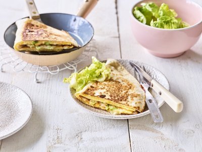 Een krokante tortilla ligt in een bordje klaar om aan te vallen. Hij is gevuld met een omelet, passendale kaas, gandaham en lente-ui. De tweede quesadilla ligt nog in de pan en staat ook op de tafel. 