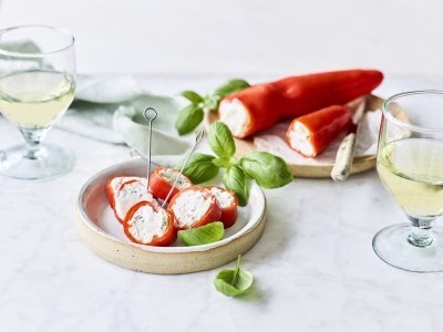 Een opgevulde puntpaprika met roomkaas en kruiden ligt op een houten snijplankje. Er werden rolletjes van gesneden en vastgeprikt met een stokje om het aperitief in stijl te starten. Voor de garnering en de kleur voegde we nog enkele verse blaadjes basilicum toe.