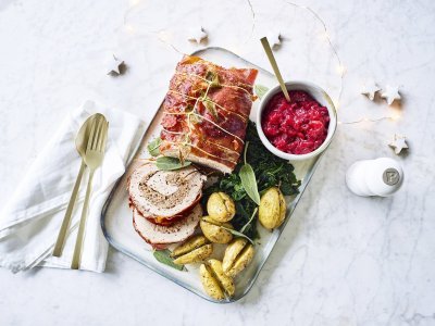 Een mooi aangesneden varkensrollade ligt op een rechthoekig dienblad. De aardappeltjes werden net niet doormidden gesneden alvorens ze de oven in gingen met een kruidentakje. De veenbessencompote staat langs de rollade die zorgt voor een intense kleur in deze feestmaaltijd. Goud bestek ligt langs het gerecht. 