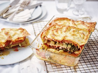 De lasagne staat op tafel in een glazen doorzichtige ovenschotel. Hij is aangesneden zodat je mooi de laagjes ziet van pasta, groenten en saus. De portie die is uitgeschept ligt op een bordje er naast.