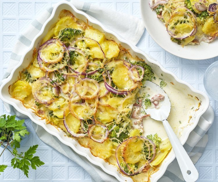 Een ovenschotel staat op tafel, waaruit al 1 hoek is uitgeschept op een bord. De ovenschotel bestaat uit krokante aardappelschijfjes, zoals een aardappelgratin, maar er is ook broccoli, rode ui en gerookte ham aan toegevoegd. Er staat een vaasje langs met een bloemetje en het tafelkleed is wit met ruitjes. 