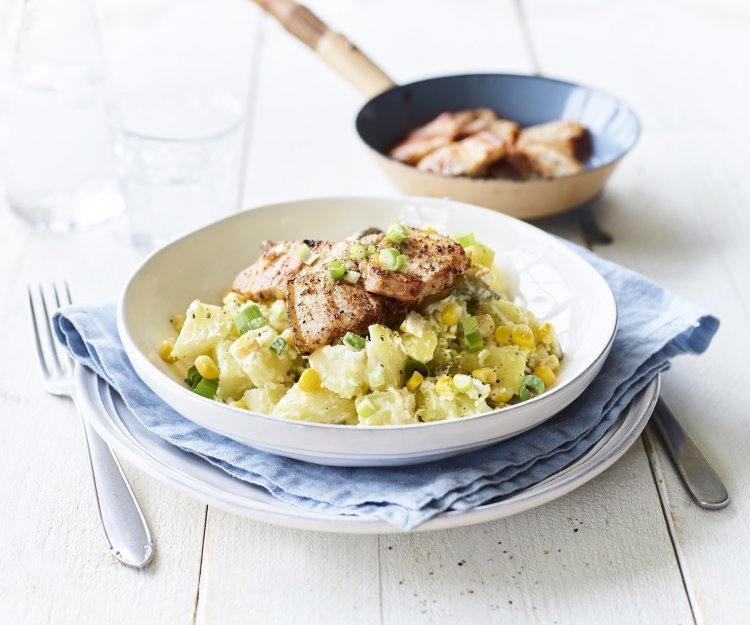 Een rijkgevulde aardappelsalade is opgediend in een diep wit bord. Hier bovenop ligt het krokant gebakken spek. De overige stukjes spek liggen nog in het bakpannetje en staat bovenaan het bord in beeld. 