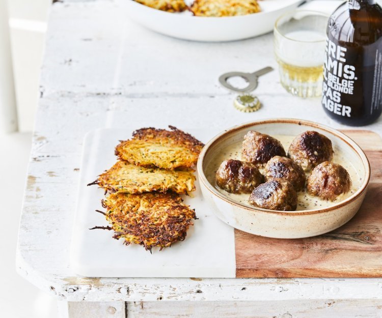 Gebakken balletjes liggen in een diep bord op de mosterdsaus, de krokante groenterösti's liggen er langs. Er staat een flesje bier op tafel met een klein glaasje.