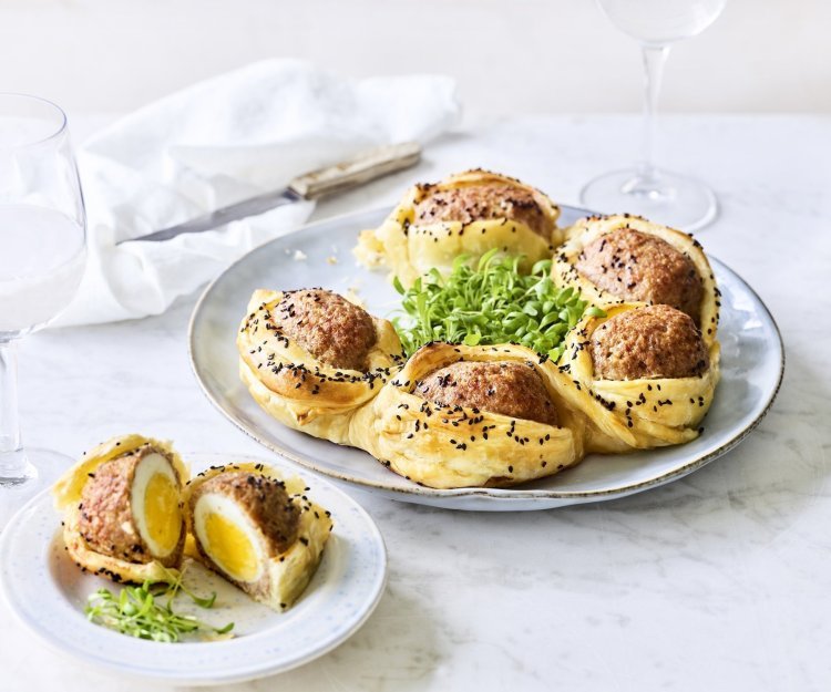 Op tafel staat een bord waarop een bladerdeegkrans ligt. Er zitten grote gehaktballen tussen gevluchten met hierin een gekookt eitje. Het bladerdeeg is afgewerkt met zwarte sesamzaadjes wat zorgt voor een leuke spikkeling.