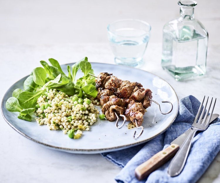 Op een lichtblauw groot bord liggen 3 gegrilde brochettes, we serveren ze met een couscoussalade van erwtjes en verse kruiden. Naast het bord ligt een blauwe servette met bestek langs. Er staat ook een glaasje water bovenaan het bord met een flesje water naast.  