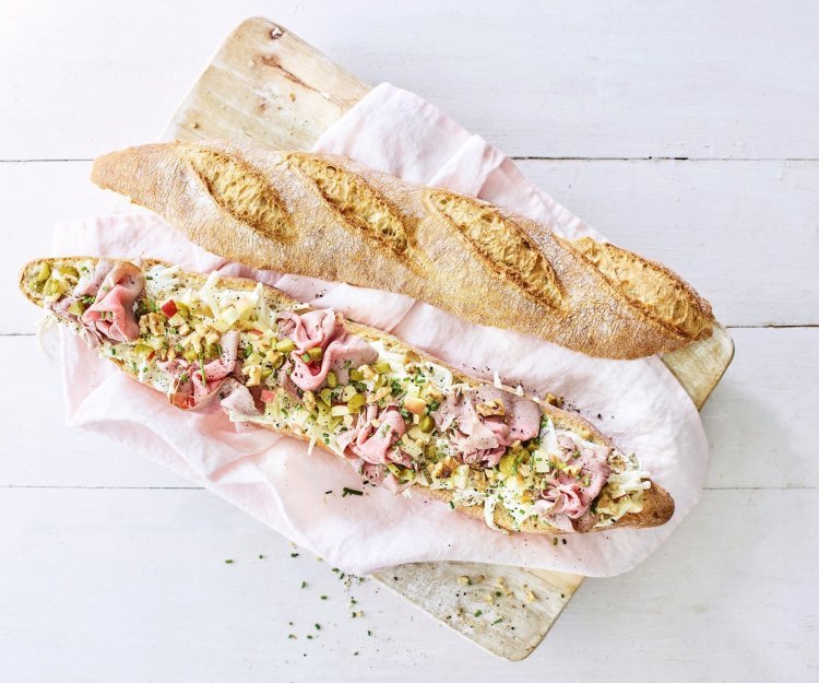 Een groot stokbrood ligt in de lengte opengesneden op een houten plank. Het broodje is belegd met een slaatje van knolselder, plukjes rosbief, stukjes appel en walnoten.