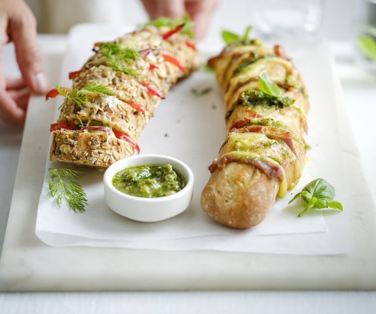 Twee broodjes liggen op een snijplank. Je ziet dat het twee versies zijn, nl een broodje met groene pesto, sneetjes ham en kaas. En dan nog een versie met roomkaas, paprika en rode ui. Extra pesto staat in een potje bij de broodjes, voor wie wat extra wil. Een vrouw zet de plateau of snijplank op tafel, haar handen zijn nog in beeld. 