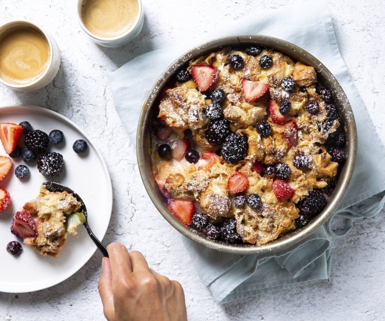 Homemade broodpudding met rabarber en zomerbessen staat op tafel. Een vrouwelijke gast schept haar een portie uit om volop te genieten van dit dessert met een kopje koffie. 