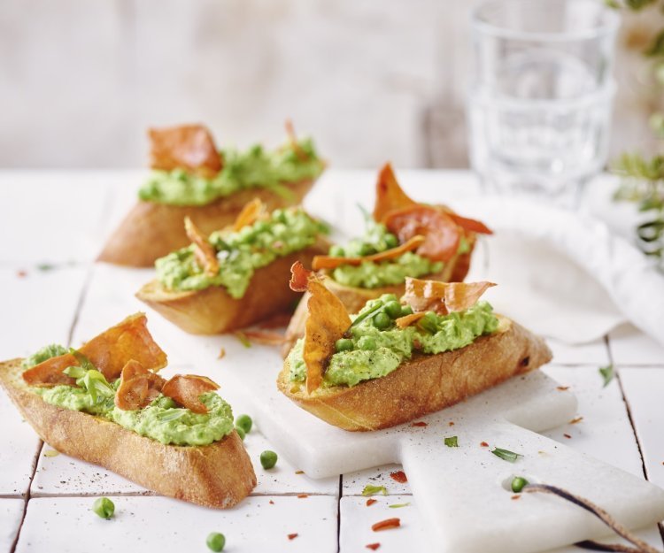 Vijf klaargemaakte bruschetta liggen op tafel. De spread van geitenkaas en erwten kleurt mooi groen, de bruchetta werd afgewerkt met een stukje krokant gebakken gedroogde ham. 