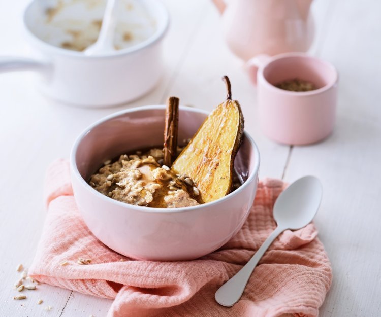 Een lichtroos ontbijtkommetje staat op tafel, met hierin de kruidige chai porridge met een halve peer en een kaneelstokje. Er is ook een roze theepot met bijpassend kopje.