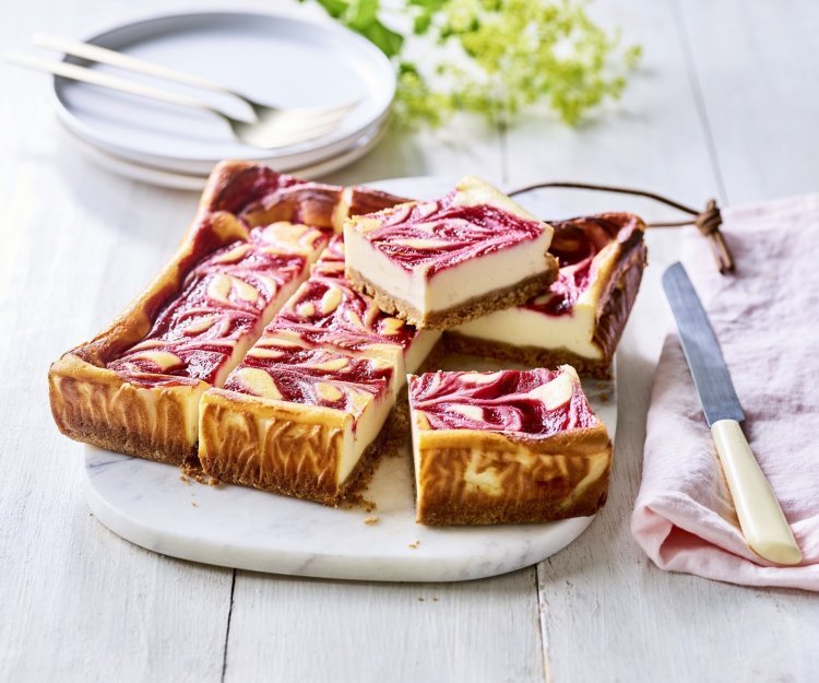 Een gebakken kaastaart staat op een marmere snijplank, het is wel een speciale cheesecake. Nl, de bovenzijde bevat frambozencoulis, waardoor er bovenaan een marmerpatroon is van de kaas en framboosjes. Echt een pareltje om naar te kijken. De taart is al aangesneden om onmiddelijk aan te vallen. 