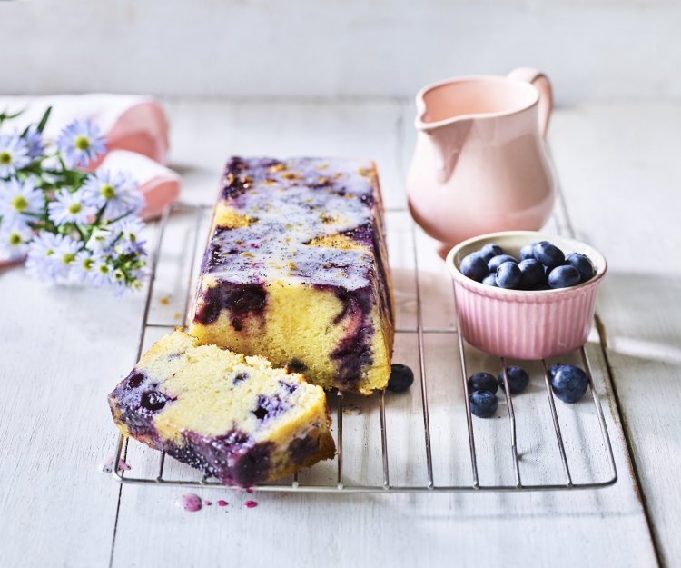 Op een bakrooster ligt de citroencake af te koelen, hij heeft een prachtige kleur door de blauwe bessen die tinten van paars achterlieten in de smeuige cake. Er staat nog een extra portie blauwe bessen naast voor extra fruitplezier.