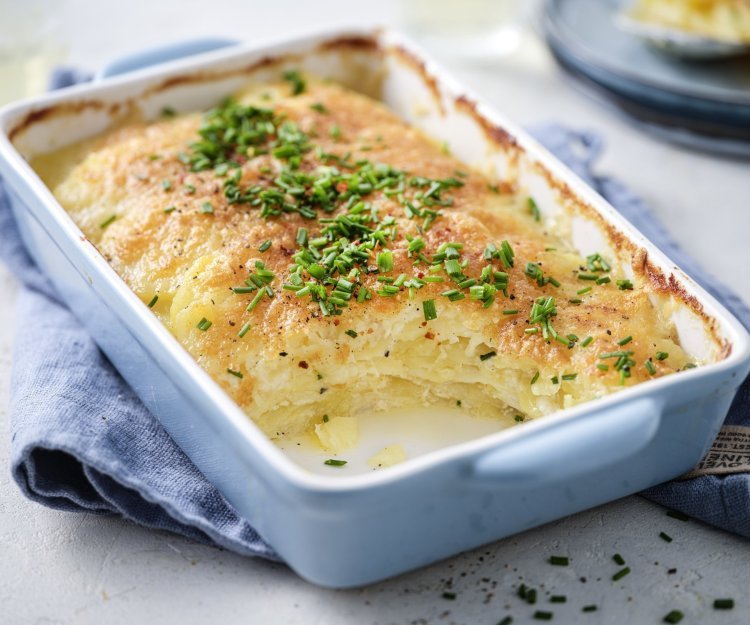 De ovenschotel met gratin staat op tafel, hij heeft een lichtblauwe kleur. De aardappelschijfjes zijn lekker crispy gebakken, doordat we ze in de oven klaarmaken met minder room. De ovenschotel is afgewerkt met verse fijngehakte bieslook.