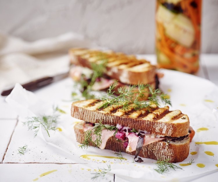 Twee croques liggen op het aanrecht in de keuken op een wit boterpapiertje. In de achtergrond staan de gepekelde groentjes (wortel en komkommer) in een bokaal. De tosti's zijn mooi gegrild in streepjes.