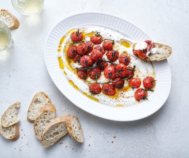 Een groot ovaal bord is gevuld met de romige yoghurt waarop de dieprode kerstomaatjes bovenop liggen. Er liggen enkele sneetjes stokbrood naast om te dippen. Er staan ook twee glaasjes witte wijn in beeld. 