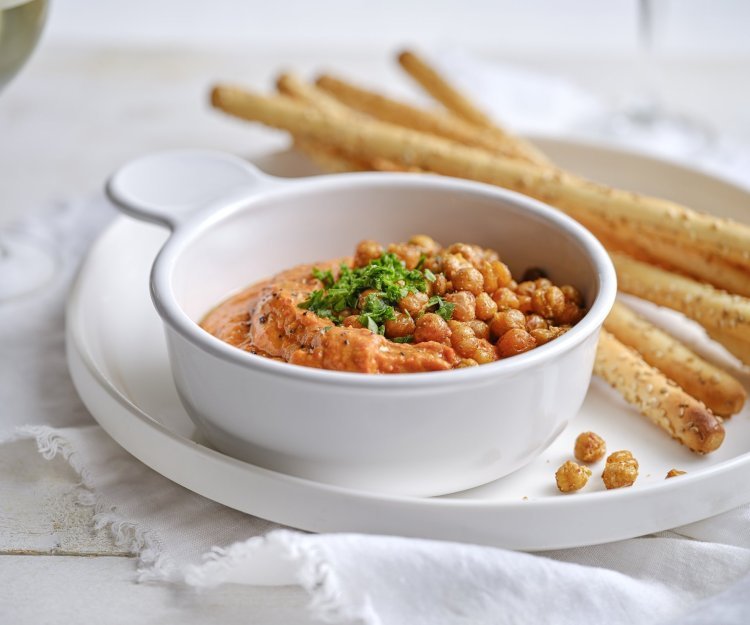 Een wit porseleine potje is gevuld met een oranjekleurige tapenade. Het is er eentje van zoete puntpaprika met hierop geroosterde kikkererwten. Er liggen broodstengels langs om te dippen. 