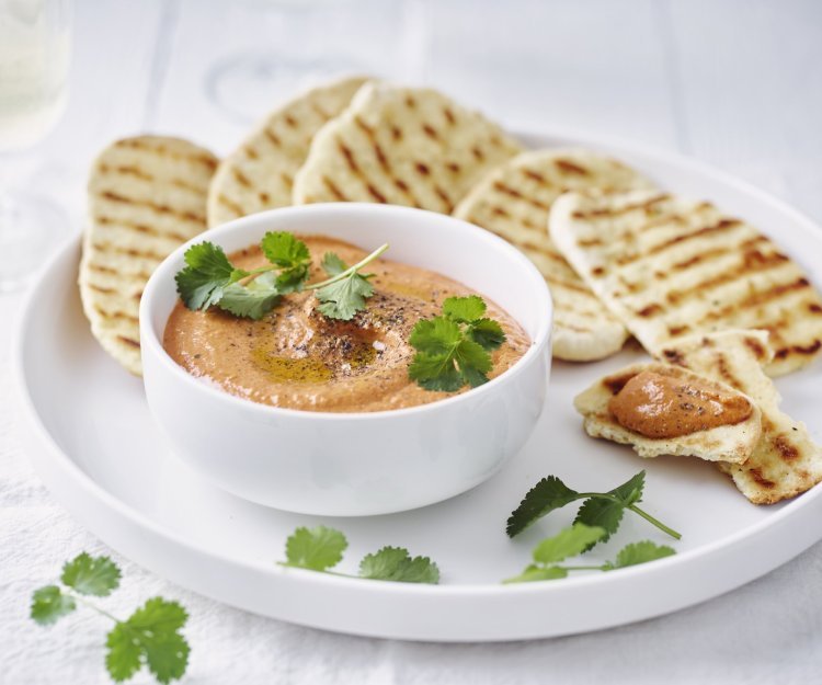 Homemade paprikadip zit in een wit schaaltjes. Het naanbrood ligt er langs op een wit bord. Het gercht is afgewerkt met korianderblaadjes.