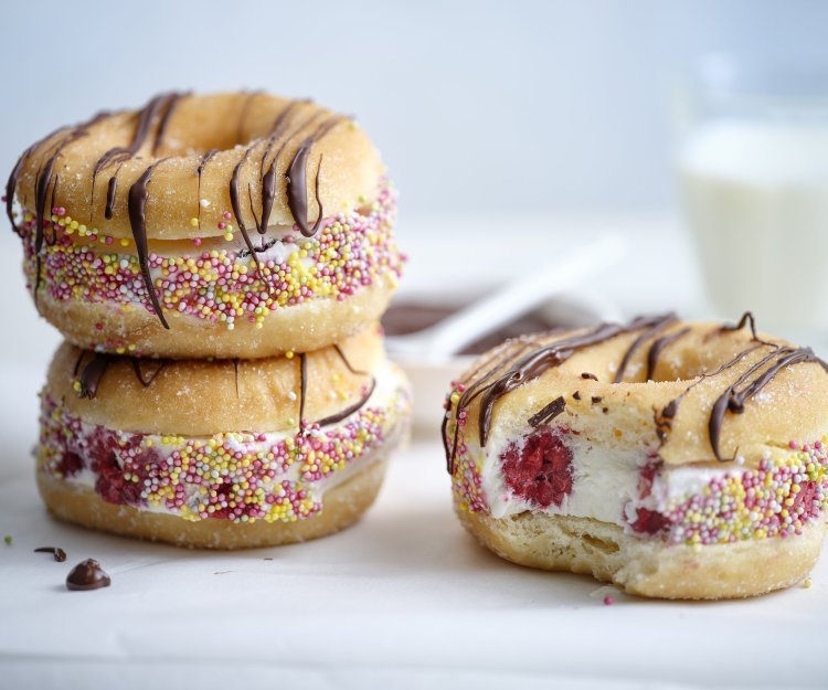 Drie donuts liggen op een witte stenen snijplank. De donuts zijn doormidden gesneden en gevuld met yoghurtijs met rode framboosjes, gerold door kleurrijke sprinkles en afgewerkt met gesmolten chocolade. In de achtergrond staat nog een glaasje melk.