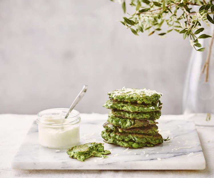 Enkele klaargemaakte fritters liggen gestapeld op een marmere snijplank. De fritters hebben een mooie groene kleur, er naast staat een glazen potje met de citroenmascarpone om te benuttigen als dip. 