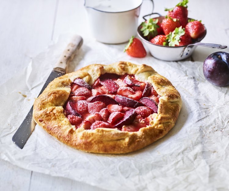 Een klaargemaakte galette staat op een houten witte tafel. Hij is prachtig goudgeel gebakken en het fruit oogt zoet-rode tinten. Er liggen nog wat aardbeien langs, alsook een paarse pruim.