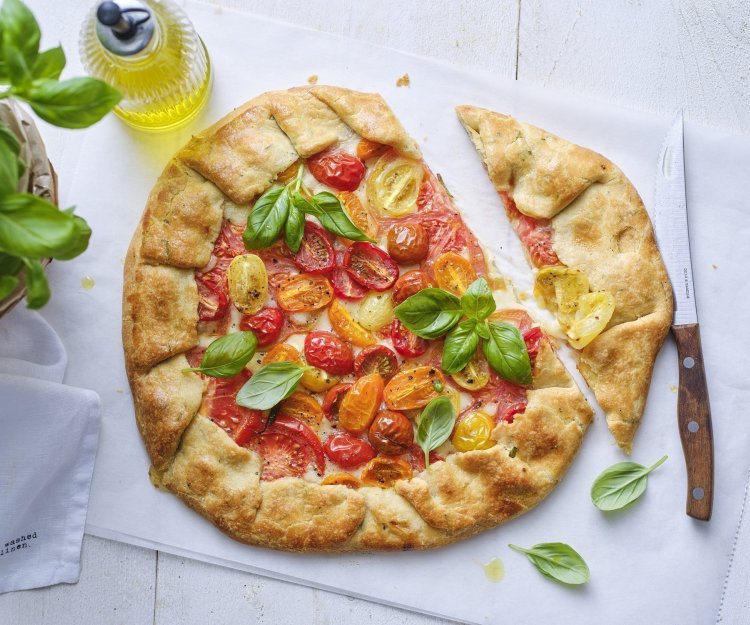 De galette van tomaten ligt op de tafel op een wit boterpapiertje. Hij is mooi gebakken en er is een stukje van gesneden. Er staat een basilicumplantje langs, alsook een flesje gouden olijfolie. 