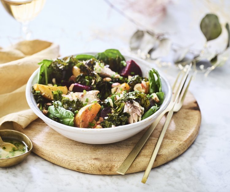 Een salade van bietjes, boerenkool en spinazie afgewerkt met noten en makreel. Deze feestsalade staat op een houten plank met de dressing in een apart gouden potje.