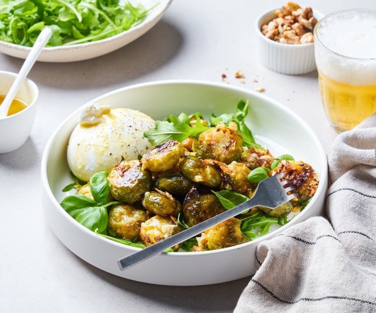 In een diep wit bord liggen de geroosterde spruitjes op een bedje van rucola en basilicum. Hierlangs ligt de burrata. Er is al wat vinaigrette gesprenkeld op de groenten en burrata.