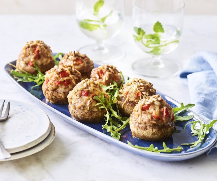 Op een blauw schaaltje liggen gevulde champignons. Ze zijn geserveerd met enkele blaadjes rucola en een stappeltje bordjes met mini-vorkjes om het hapje proper te eten. 