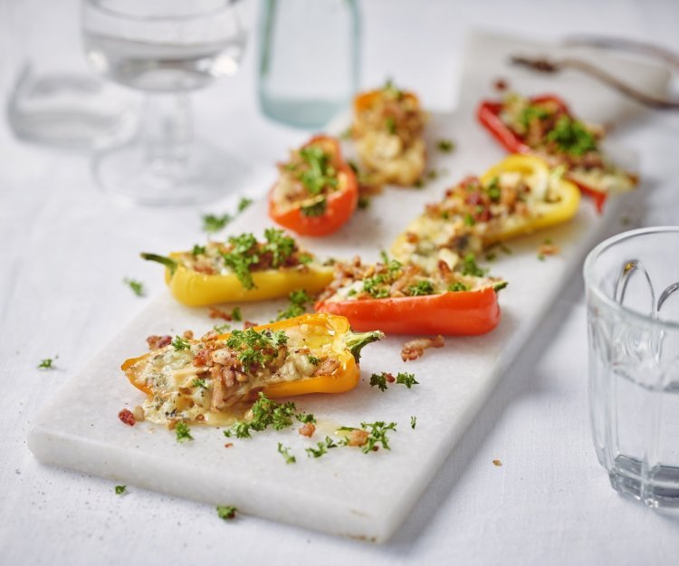 Gele en oranje snackpaprika's liggen op een witte stenen snijplank. De paprika's zijn gevuld met kaas, noten en crumble van spek. Ze zijn afgewerkt met verse peterselie. 