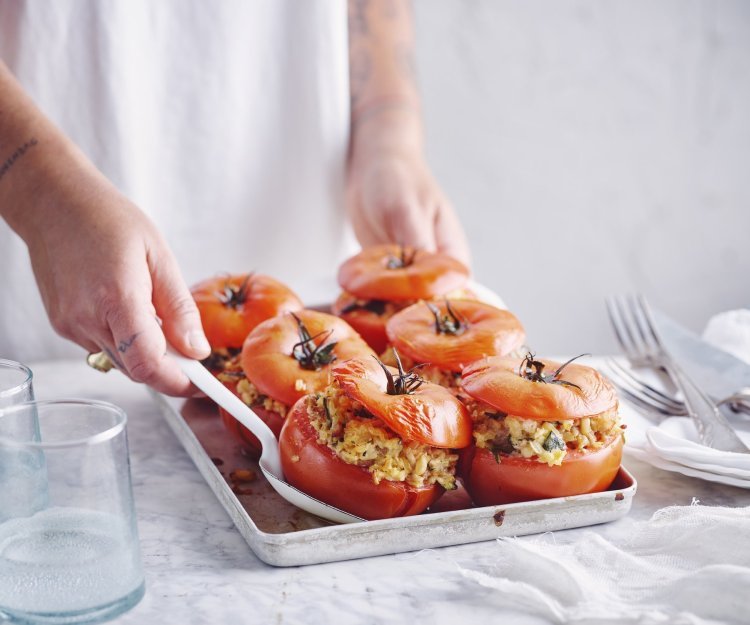 Zes gevulde rode tomaten in een ovenschaal zijn aan tafel gebracht door een vrouw in witte t-shirt. Ze schept alvast eentje op voor zichzelf.