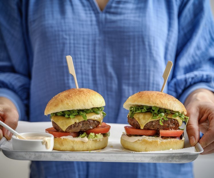 Een vrouw staat in beeld met haar romp draagt een plateau met hierop twee hamburgers. De hamburgerbroodjes hebben een mooi goudgele kleur, ze werden gemaakt met aardappelpuree in het deeg. Verder zijn de burger belgd met een schijfje vlees, kaas, tomaat, sla en saus natuurlijk. 