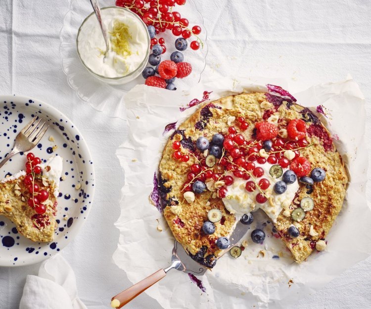 Een aangesneden taart staat op tafel met een wit laken er op. De kleuren van het rood fruit geven hierdoor nog een mooier zomers kleur. Naast de havermouttaart staat een bordje met blauwe spikkels en hier het stukje taart erop en een glaasje met de topping van de platte kaas en citroenzeste. 