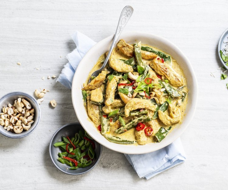 Er staat een grote witte serveerschaal op tafel gevuld met de goudgele kip curry. Je ziet de groene peultjes en rode chili duidelijk. Langs het gerecht staan nog extra noten, gesneden lente-uitjes en een bordje rijst. 