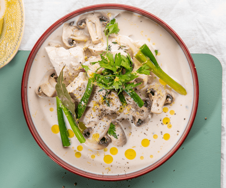 Kip gegaard in melk staat op tafel in een diep beige bord met bordeau randje. Er liggen lente-uitjes en champignons bij. Het bordje staat op een blauwgroene onderlegger. 