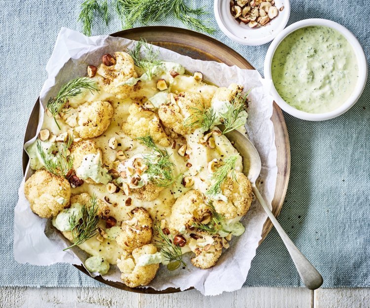 Puree van knolselder is uitgeschept op een groot lichtbruin bord. Hierop zijn de geroosterde bloemkoolroosjes geserveerd met wat drizzle van de groene kruidendressing, noten en verse dille. De overige kruidendressing is in een witte potje en staat langs het bruine bord, samen met nog wat gehakte noten in een ander potje. Alles staat op een een plaid in een blauwgrijze kleur.