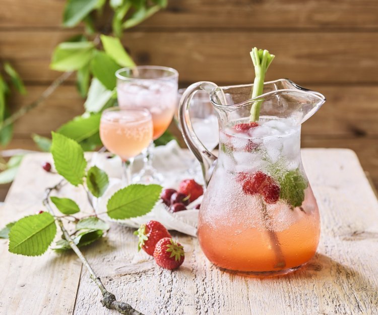 Er staat een karaf op een houten bruine buitentafel. Hij is gevuld met homemade limonade van rabarber en aardbeien. Er zitten ijsblokjes, muntblaadjes, aardbeien en een tak rabarber in de karaf. Er zijn twee glaasjes uitgeschonken. 