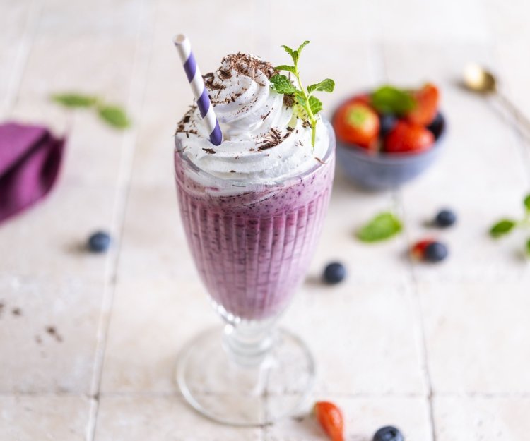 Een heerlijke grote milkshake staat op het aanrecht te wachten. Hij is mooi paars van kleur door de blauwe besjes gemixt met aardbeien. Er liggen enkele bessen op het aanrecht. De milkshake werd afgewerkt met een toef slagroom, muntblaadje en chocoladeshilfers. Ten aanval! 