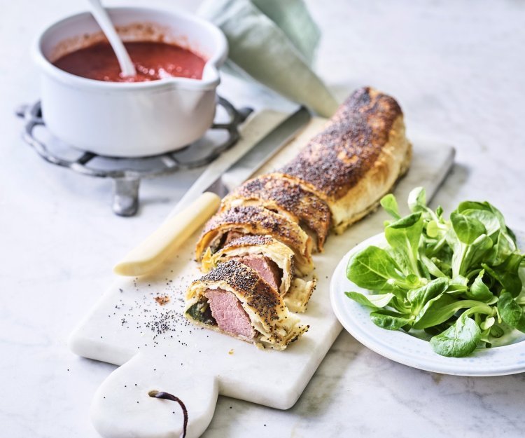 Een rol bladerdeeg ligt op een marmere snijplank. Het bladerdeeg is al aangesneden en zo zie je de spinazie en het vlees zitten. Wij serveren hem in kleine stukjes, net zoals een sushi, met homemade tomatensaus. 