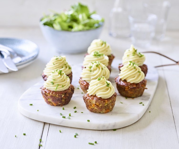 Gehaktbroodjes die klaargemaakt zijn in een muffinvorm staan recht op een marmer snijplankje. Ze zijn afgewerkt met een toefje aardappelpuree dat er werd opgespoten. Ze zijn een feest om naar te kijken.