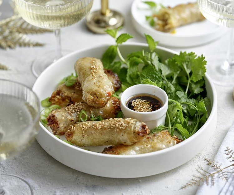 In een wit bordje liggen klaargemaakte miniloempia's, ideaal fingerfood. Ze liggen op een bedje verse kruiden en werden geserveerd met een dipsausje op basis van sojasaus. De tafel is gezet in feeststemming met mooie glaasjes en stoffen servetten.